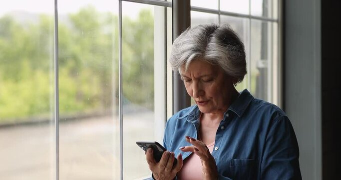 Grey-haired female standing indoor near window with smartphone modern device, swiping, searching goods discuss purchase distantly while talk on speakerphone on mobile phone. Modern tech usage concept