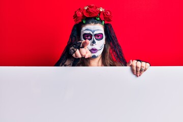 Young woman wearing day of the dead costume holding blank empty banner pointing with finger to the camera and to you, confident gesture looking serious