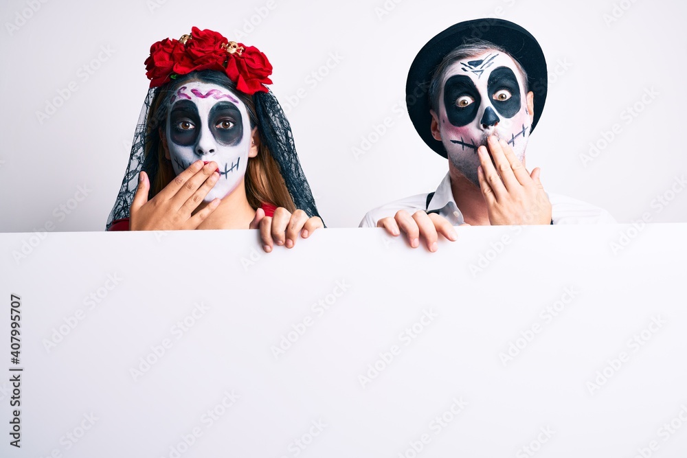 Sticker couple wearing day of the dead costume holding blank empty banner covering mouth with hand, shocked 