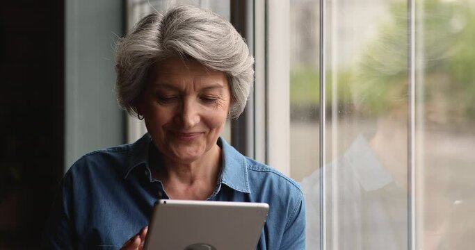 Attractive grey-haired older female standing lean on panoramic window indoor use tablet device, make delivery order on-line, scrolling news feed, enjoy easy comfort modern tech, internet usage concept