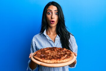 Beautiful hispanic woman holding tasty pepperoni pizza afraid and shocked with surprise and amazed...