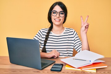 Brunette teenager girl working at the office with laptop smiling with happy face winking at the camera doing victory sign. number two.