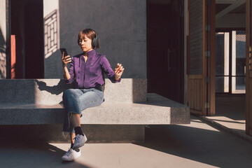 Happy young asian woman listening to music with headphones via smartphone and having fun while sitting side the street.