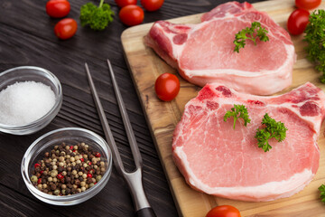 Meat steaks. Raw pork or beef ribeye meat with bone. Ingredients for barbecue. Cherry tomatoes, colored pepper, sea salt and parsley on black wooden background. Veal chunk. Food photography.