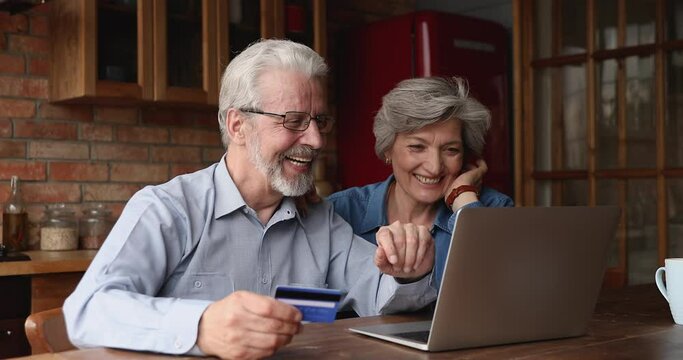 Smiling old couple sit in kitchen buying online using laptop and credit card, e-commerce e-banking services satisfied clients, safe and secure payment, successful instant money spend distantly concept