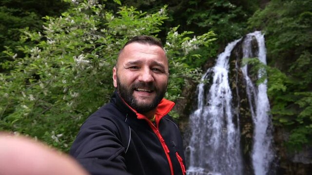 Young, happy man taking selfie photo by beautiful waterfall, slow motion