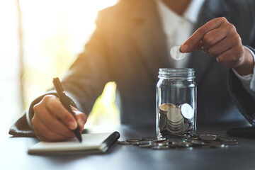Closeup image of a woman putting coins in a glass jar and taking note for saving money and financial concept