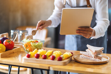 A woman following recipe on digital tablet while cooking salad and sandwich in the kitchen, online learning cooking class concept