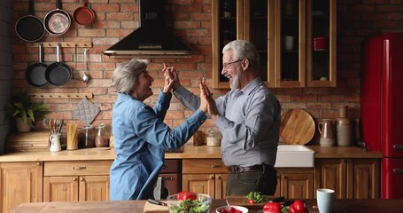 Happy active elderly spouses distracted from vegetable salad preparation dancing in modern cozy kitchen. Aged couple celebrate life event anniversary listening music moving enjoy romantic date concept - Powered by Adobe