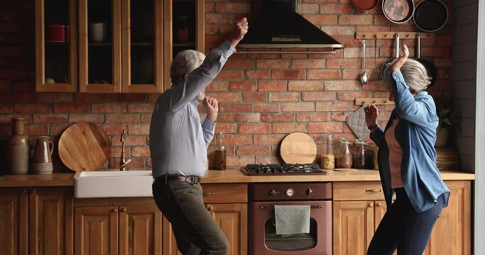 Elderly wife dancing spinning hold hands with husband couple listen music having fun together in cozy kitchen. Romantic date, relations of older generation people. Carefree, active old spouses concept