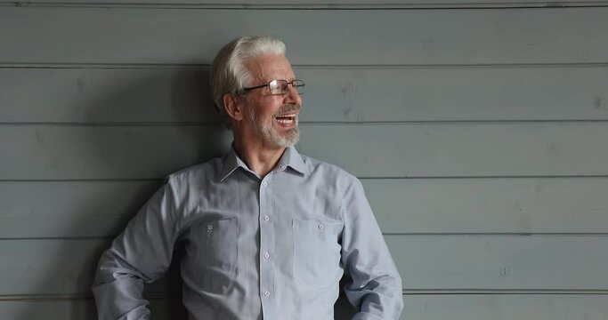 Half-length portrait laughing older man in glasses posing in studio. Grey-haired bearded male standing indoor lean on wooden wall background looking away into distance giggling feels healthy and happy