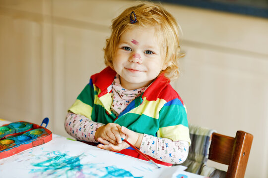 Cute adorable baby girl learning painting with water colors. Little toddler child drawing at home, using colorful brushes. Healthy happy daughter experimenting with colors, water at home or nursery.