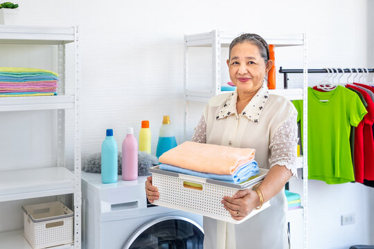 Happy smiling Asian senior elderly female woman carrying clothe basket doing laundry with washing machine, house cleaning and housekeeping, looking at camera