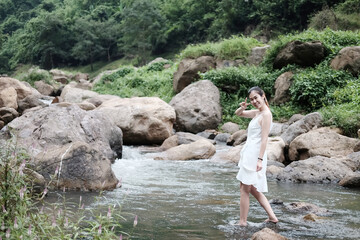Smiling Beautiful asian female in white dress is standing on the rock and stream in the tropical forest.