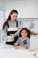 Smiling Asian mother and little asian girl child is Kneading the dough for baking bakery on wooden table together in kitchen. Homemade pastry for bread. Family love and Homeschool Concept.