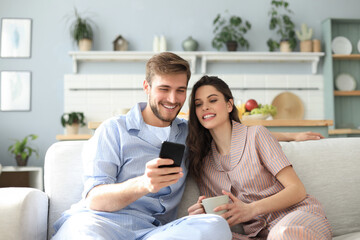 Young couple in pajamas watching online content in a smart phone sitting on a sofa at home in the...