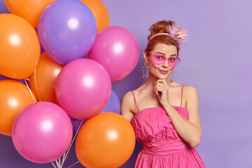 Obraz premium People celebration and holidays concept. Fashionable nineties woman looks gladfully at camera wears clothes in vintage style prepares for party poses with balloons isolated over purple background