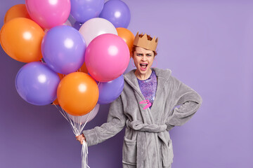 Fototapeta na wymiar Annoyed young woman in soft robe and handmade paper crown has birthday party celebrates holiday holds bunch of multicolored inflated balloons isolated over purple background. Celebration concept