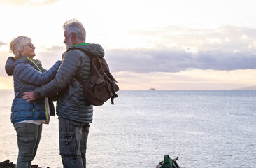 Sporty elderly couple on hike on ocean cliffs. On the horizon the profile of an island and a ship. Cloudy sky at sunset