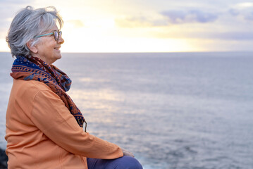 Fototapeta na wymiar Relaxed senior grey-haired woman enjoying the freedom and the horizon over water. Sitting on the cliff in front to the sea in a winter cloudy day