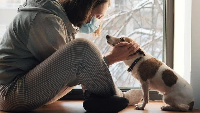 Woman sitting in medical mask at the window with a shaggy dog. Self-isolation concept virus covid 19 infection doomsda coronavirus