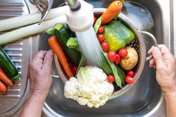 Two female hands hold a colander full of fresh vegetables from the garden under the jet of water. Carrot, zucchini, potatoes, pepper, cauliflower, tomatoes and green been