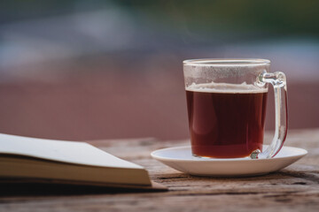 A cup of hot morning coffee on wooden table with a diary notebook at the balcony of the sleeping room to welcome sunshine in the morning, a glass of black coffee with a notebook on the wooden table