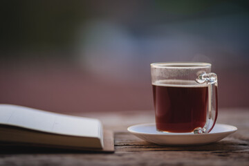 A cup of hot morning coffee on wooden table with a diary notebook at the balcony of the sleeping room to welcome sunshine in the morning, a glass of black coffee with a notebook on the wooden table