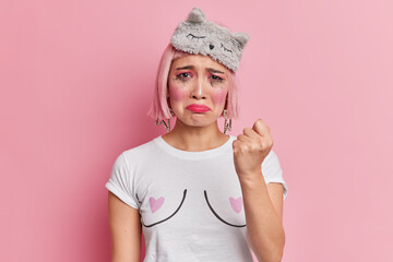 Unhappy Asian woman clenches fist and promises to revenge for being offended has spoiled makeup after crying wears sleepmask and casual t shirt isolated over pink background. I will show you