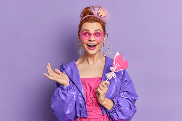 Retro fashion model dressed in nineties style has happy expression nostalgia holds delicious candy poses over vivid purple background. Fashion trends. Redhead girl in trendy sunglasses purple jacket