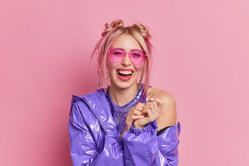Happy European woman with trendy hairstyle giggles positively has fun at disco with friends wears bright fashionable clothes prefers style from ninenties smiles broadly isolated over pink background