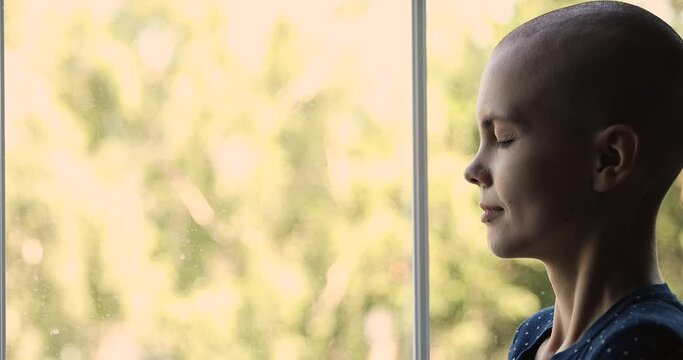 Close up side profile view face of thoughtful sad female cancer patient. Young pensive bald after chemotherapy woman standing indoor alone look out window thinking about oncology disease feel hopeless