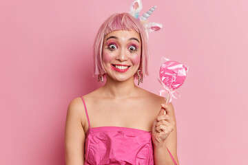 Obraz na płótnie Canvas Happy pinup girl with toothy smile has leaked makeup after crying being calmed by getting delicious sweet candy on stick dresses for carnival party isolated over pink background. Positive Asian woman