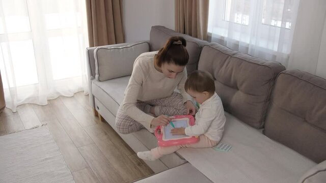 Caring young mother or nanny helping her cute daughter teach the baby, draw a picture with pencils, lying on the sofa in the living room, learn creativity at home. High quality 4k footage