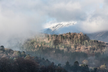 Dramatic landscape image looking across Derwentwater in Lake District towards Catbells snowcapped mountain with thick fog rolling through valley