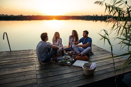 Group of friends having fun on picnic near a lake, sitting on pier eating and drinking wine.