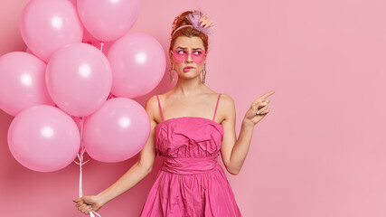Obraz na płótnie Canvas Puzzled redhead young woman dressed in glamour outfit wears everything pink poses on hens party with inflated balloons points at empty space bites lips shows place for your advertising content
