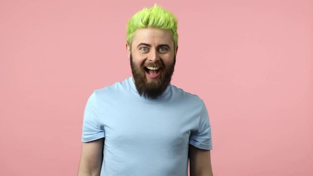 Extremely positive bearded man with dyed bright green hair suddenly appearing and waving hand looking at camera, greeting and welcoming you. Indoor studio shot isolated on pink background