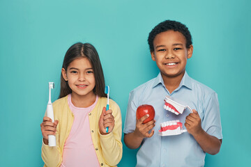Asian girl and African American boy advertising oral hygiene and brushes teeth. Kids holding manual and electric ultrasound toothbrushes