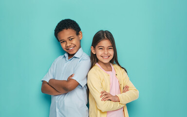 Asian girl and African American boy standing back to back with arms crossed on a turquoise...