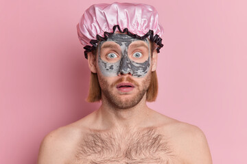 Portrait of suprised bearded man stares shocked at camera wears shower hat applies nourishing clay mask for rejuvenation and reducing wrinkles stands with bare shoulders isolated over rosy background