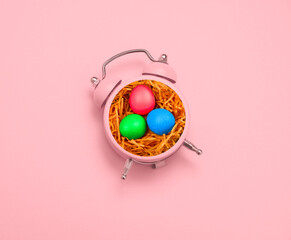 Top view creative minimal art collage alarm clock and bird nest with painted eggs on a pastel pink...
