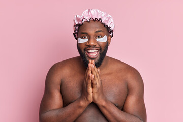 Happy dark skinned man keeps palms together has good mood applies moisturising patches under eyes wears shower hat stands with naked body isolated over pink background. Topless Afro American guy