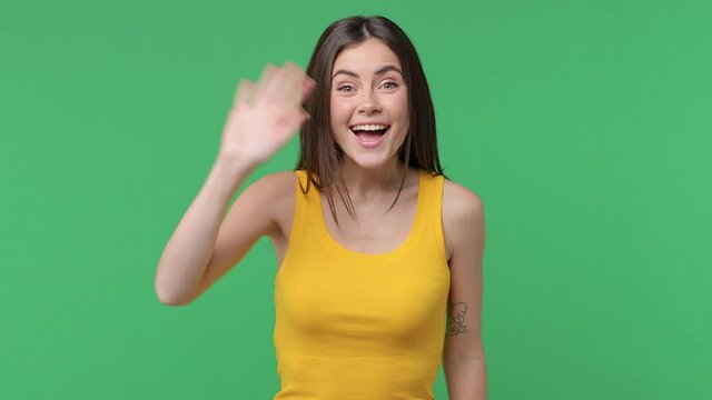 Excited cheerful brunette young woman 20s years old in yellow basic tank top posing isolated on green background in studio. People lifestyle concept. Waving and greeting with hand as notices someone