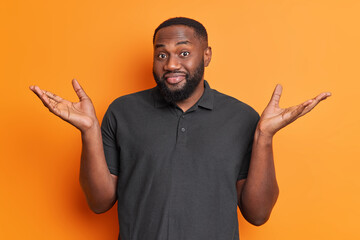 Dark skinned clueless man with beard spreads palms shrugs shoulders looks unaware faces difficult choice dressed in casual black t shirt isolated over vivid orange background. Human perception - obrazy, fototapety, plakaty