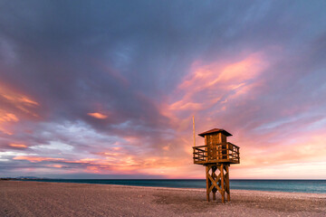 Torre de vigilancia en una playa del Mediterraneo que destaca sobre un cielo con nubes teñidas de...
