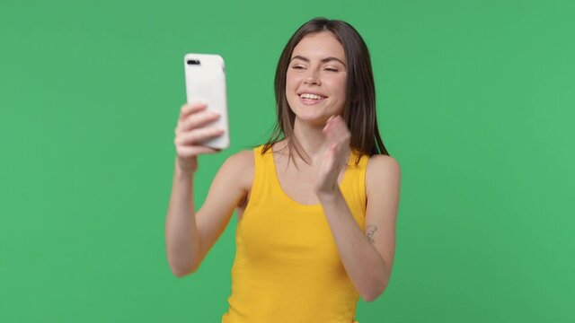 Funny young woman 20s in yellow tank top isolated on green background studio. People lifestyle concept. Doing selfie shot on mobile phone greeting hand showing victory sign blowing sending air kiss