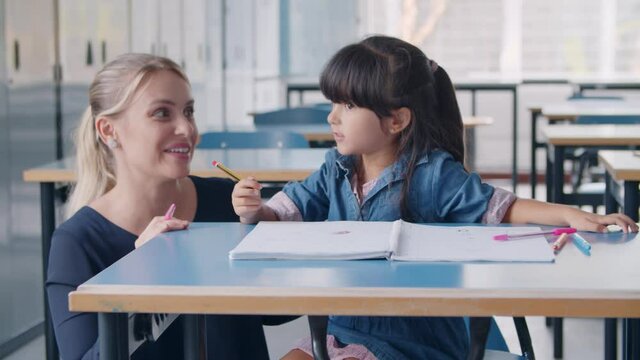 Happy female school teacher and Latin schoolgirl discussing task. Primary schoolkid holding pen, sitting at desk with copybook and speaking. Education or teaching concept