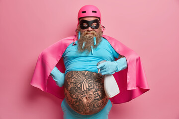 Serious powerful superman holds detergent ready for cleaning room wears helmet mask undersized t shirt claok and rubber gloves has big tattooed belly busy doing house chores isolated over pink wall