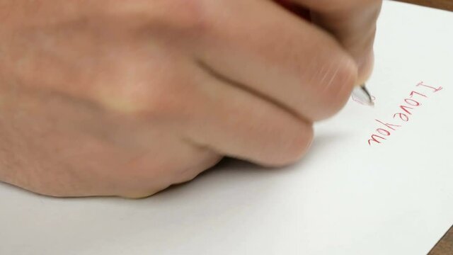 Close-up of a young man's hand he writes on paper the words I love you with a red pen.